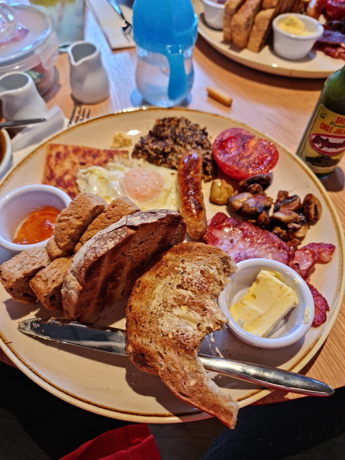 A full Scottish breakfast on a speckled plate: tattie scone, haggis, a fried egg, a sausage, grilled tomato, mushrooms, bacon, brown toast and butter, with a pot of relish.