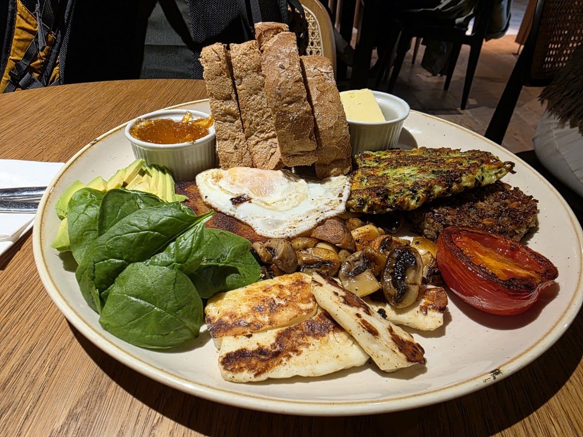 A veggie brunch plate: grilled halloumi, spinach, avocado, a courgette fritter, a fried egg, mushrooms, a roasted tomato and sliced toast with butter and marmalade.