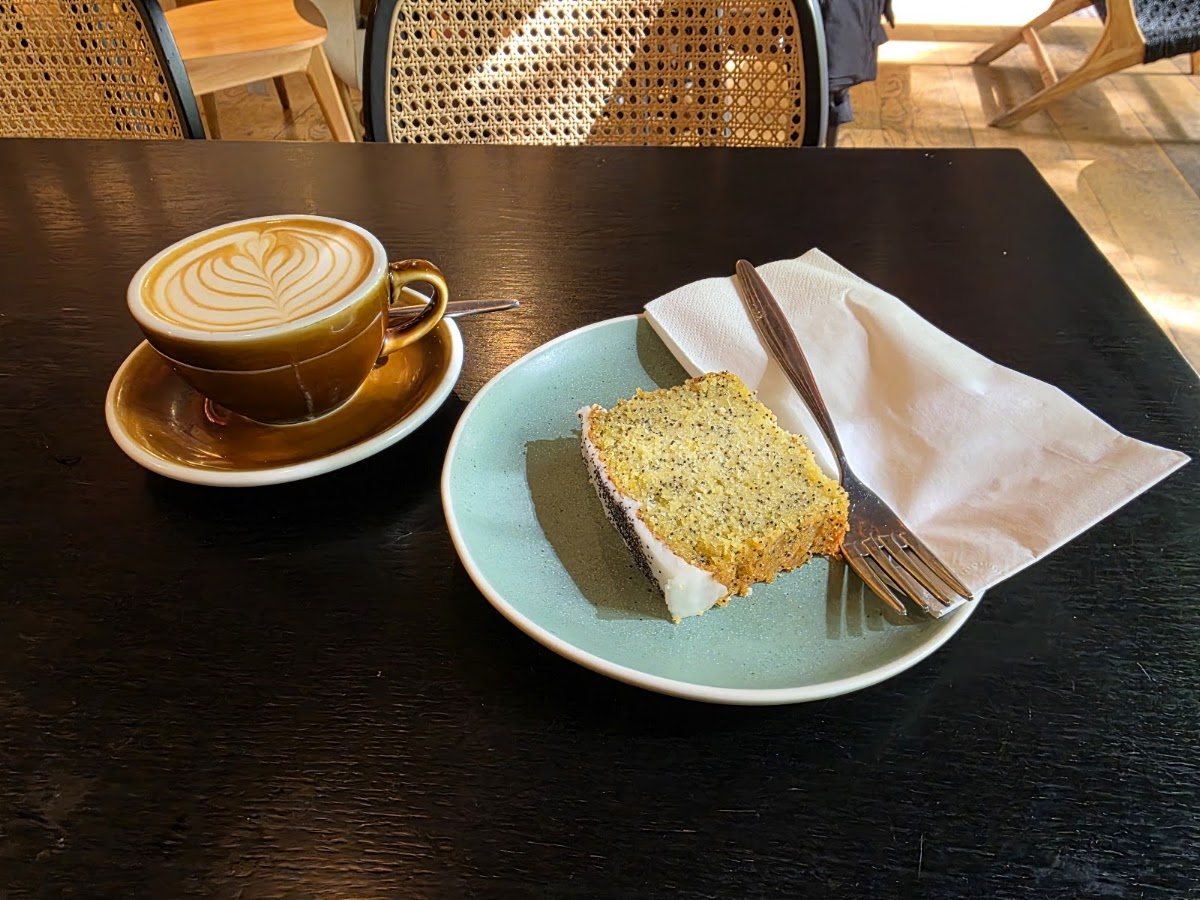A flat white with latte art in an ochre cup, beside a slice of iced lemon and poppy seed cake on a pale blue plate, on a dark table.