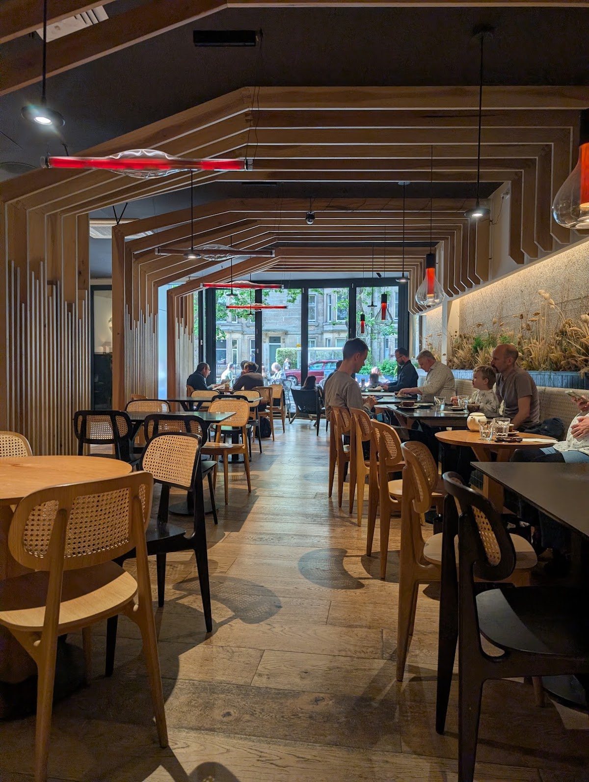 Inside Two Children Coffee: pale oak slatted frames step down the length of the room like a timber tunnel, over round wooden tables and cane-backed chairs, with red glass pendant lights and the big front windows onto Raeburn Place at the far end.